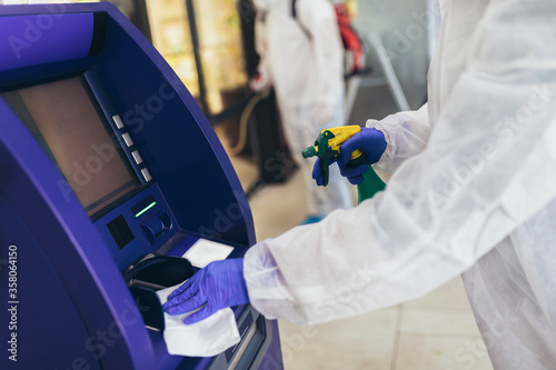 Professional workers in hazmat suits disinfecting ATM machine.