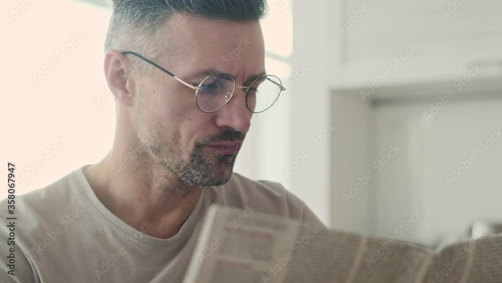 A close-up view of a serious focused man is reading a newspaper at home in the kitchen at the morning A close-up view of a serious focused man is reading a newspaper at home in the kitchen at the morning