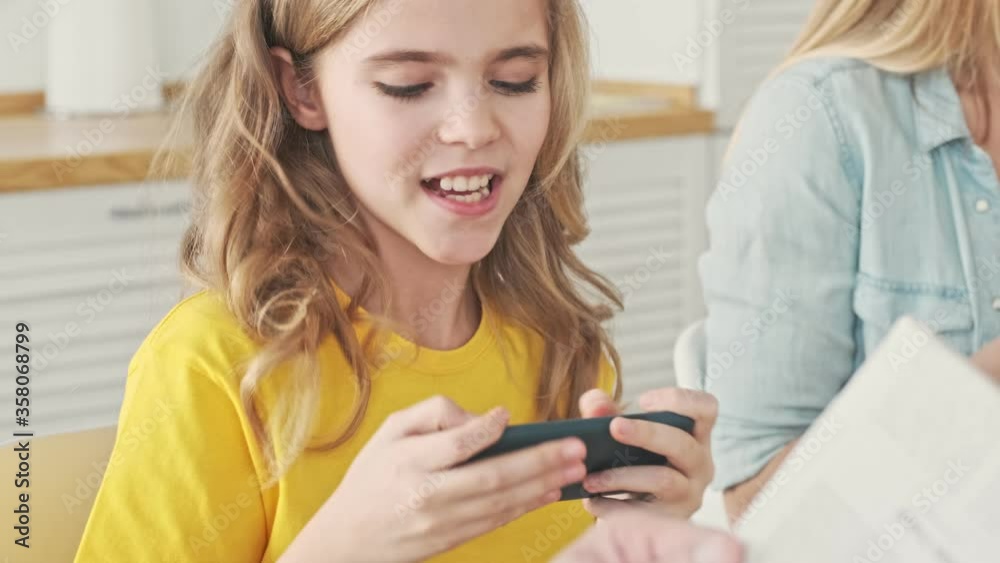 An emotional happy young girl is playing games on her smartphone holding it horizontally during breakfast at home in the kitchen at the morning An emotional happy young girl is playing games on her smartphone holding it horizontally during breakfast at home in the kitchen at the morning