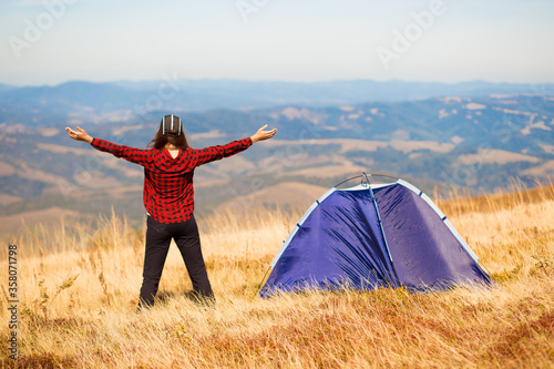Back view of woman hiking mountain trail  standing near tent with hands up  enjoying summer morning. Camping adventure vacations outdoor
