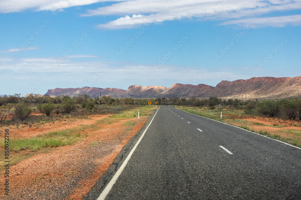 Driving towards the MacDonnell ranges. Empty road, no cars. Typical yellow Australian road signs. Green vegetation and bush on the sides. MacDonnell ranges, Northern Territory NT, Australia, Oceania