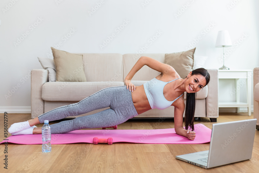 Girl training at home, doing workout and watching videos on laptop, training in living room. Beautiful woman is doing side plank exercise. She is watching video on the internet and repeating the tasks Girl training at home, doing workout and watching videos on laptop, training in living room. Beautiful woman is doing side plank exercise. She is watching video on the internet and repeating the tasks