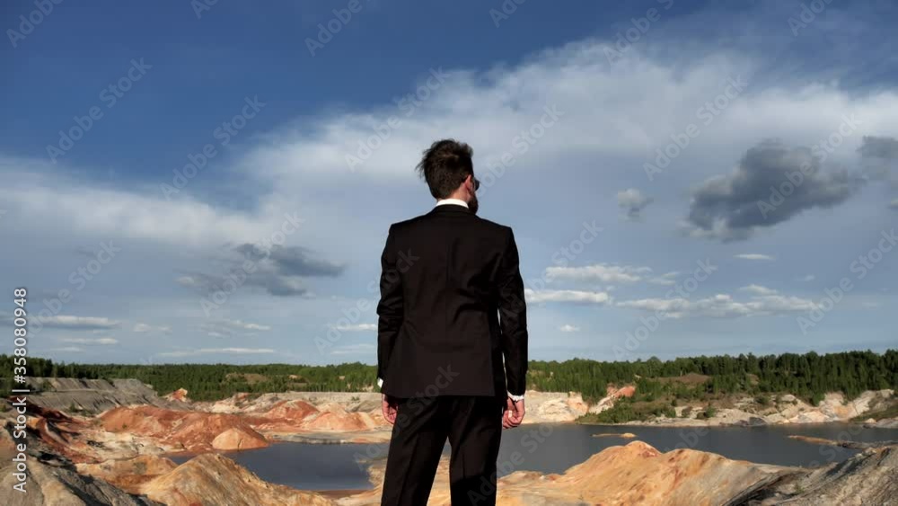 A businessman with a beard in sunglasses, a business suit stands with his back to the camera on a hill and looks into the distance on a hilly Martian landscape with a small pond, a man turns his head A businessman with a beard in sunglasses, a business suit stands with his back to the camera on a hill and looks into the distance on a hilly Martian landscape with a small pond, a man turns his head