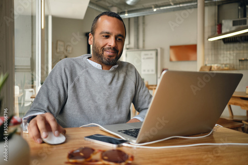 Businessman in sweater happily working on laptop during coffee break in cafe