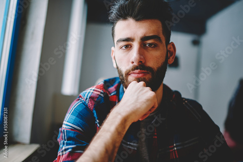 Portrait of concentrated handsome hipster guy 20 years old sitting indoors and looking in camera after course work,