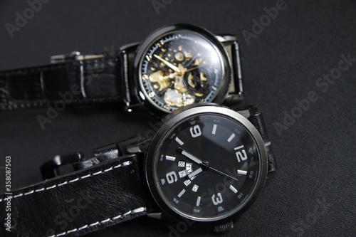 a black automatic self winding wristwatch with transparent sekeleton dial design and a black battery operated watch on black leather background