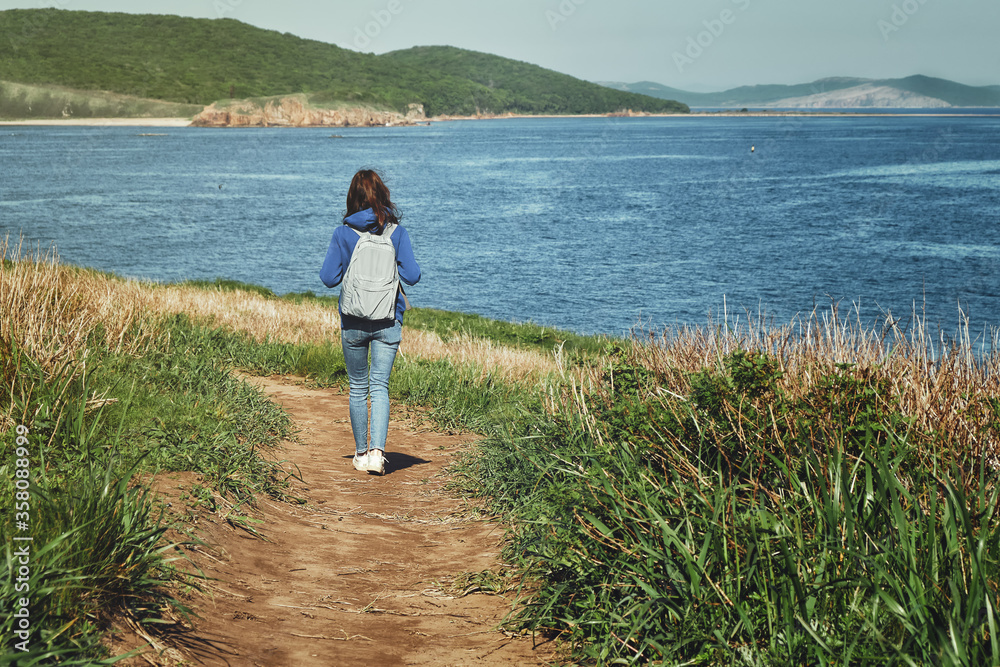back view of a young girl with long hair and backpack walks alone along a path goes along a hill above the sea with a view of the island overgrown with grass. Social distancing and travel concept