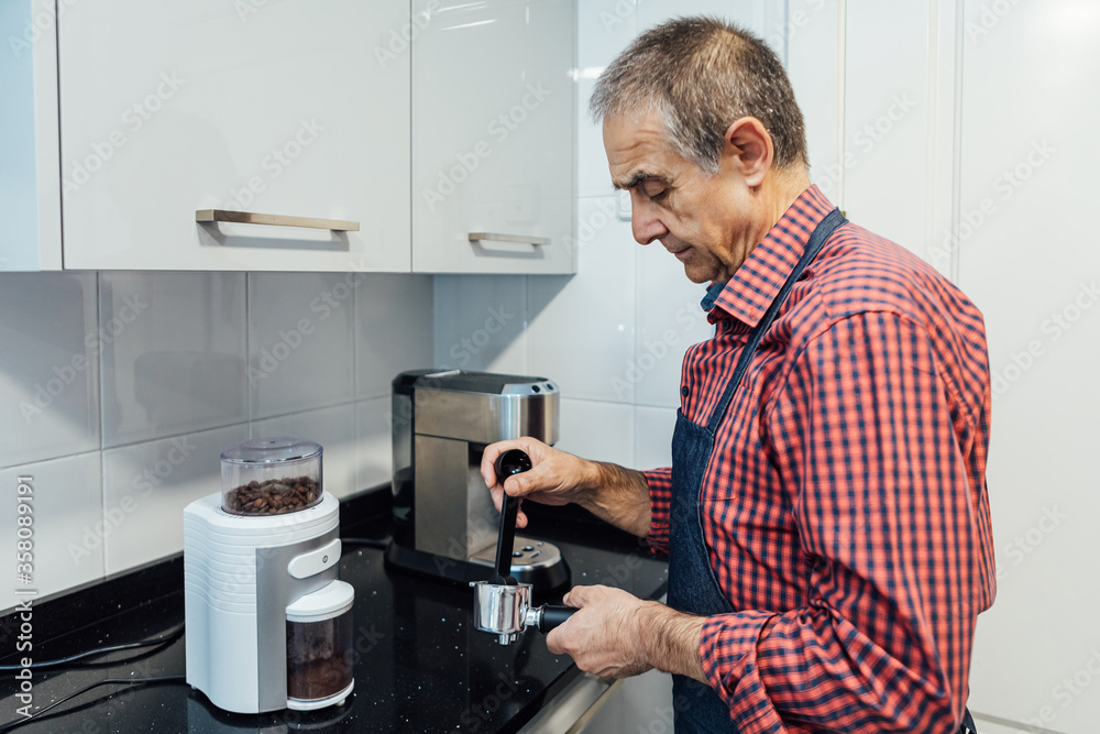 Mature man dressed in plaid shirt and apron brews espresso at home Mature man dressed in plaid shirt and apron brews espresso at home