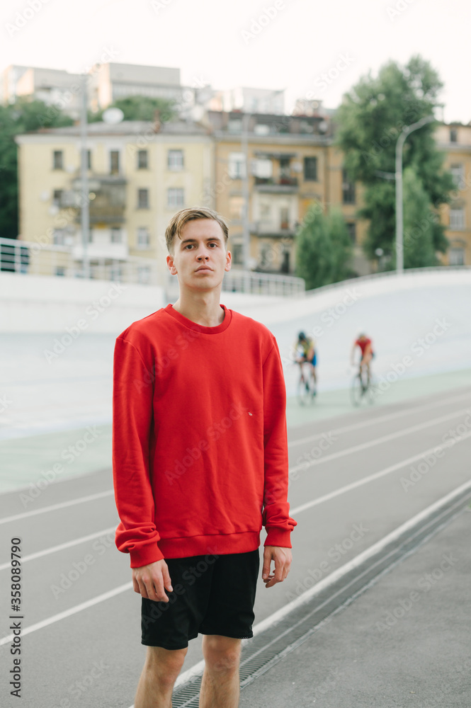 Young man in a red sweatshirt stands on a sports track on a background of velodrome and bicycle.Portrait of a cyclist without a bicycle posing for the camera on the background of the velodrome tracks Young man in a red sweatshirt stands on a sports track on a background of velodrome and bicycle.Portrait of a cyclist without a bicycle posing for the camera on the background of the velodrome tracks