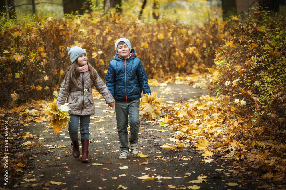 A boy and a girl walk along the path in the autumn park, they hold hands and carry a bouquet of leaves. They are wearing hats and jackets. A boy and a girl walk along the path in the autumn park, they hold hands and carry a bouquet of leaves. They are wearing hats and jackets.