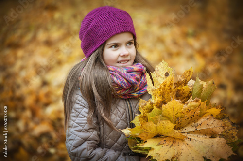 Little beautiful girl stands with a bouquet of maple orange leaves in the autumn park. She looks away and smiles. Dressed in a burgundy knitted hat  her hair loose. Close-up  soft focus.