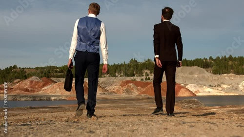 Two businessmen are walking from the camera towards the unusual Martian landscape, a man in a white shirt and vest is holding a bag in his hands, the characters are moving away from the camera.