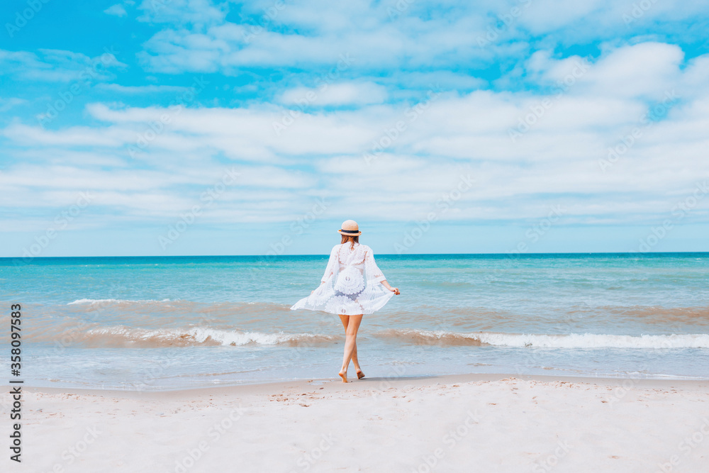 Beautiful sexy woman wearing straw hat, white swimsuit and skirt walking along the surf line on beach. Summer holidays in tropics. Copyspace. Sun protection.