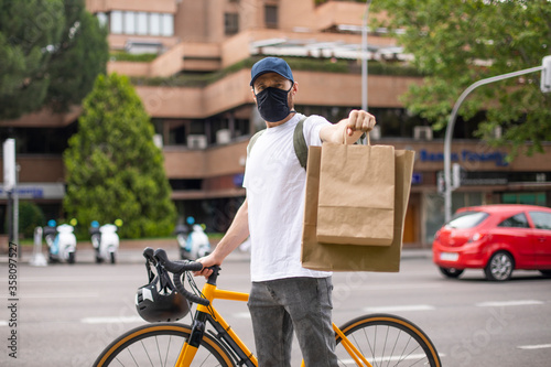 Repartidor de comida a domicilio con mascarilla en bicicleta amarilla por la ciudad con la mochila cargada de pedidos mostrando bolsas de cartón
