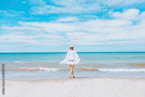 Beautiful sexy woman wearing straw hat, white swimsuit and skirt walking along the surf line on beach. Summer holidays in tropics. Copyspace. Sun protection.