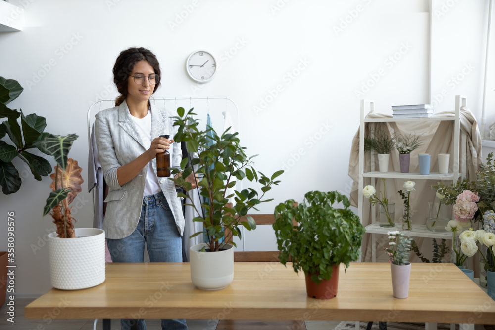 Young Caucasian woman take care of green plants in office or home spray sprinkle liquid fertilizer on leaves, millennial female watering domestic flowers, hobby, gardening, horticulture concept Young Caucasian woman take care of green plants in office or home spray sprinkle liquid fertilizer on leaves, millennial female watering domestic flowers, hobby, gardening, horticulture concept