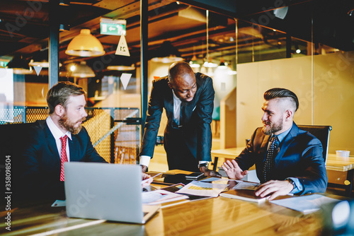 Diversity team of prosperous male entrepreneurs in formal wear discussing sponsorship of project on meeting table,experienced group of economists talking about finance planning of corporation