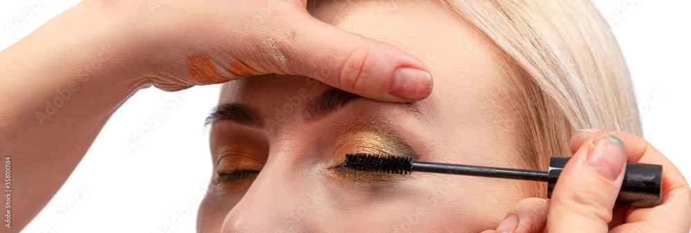 Makeup artist paints the model’s eyelashes with a black mascara brush on her eyes closed with oriental make-up and golden shadows in the studio on a white background. Banner for advertising artvizage.