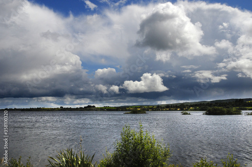 Vue panoramique sur un lac et ses îlots de terre, avec une ruine de tour médiévale en son centre. Ciel chargé et nuageux, collines, verdure et rochers. © Delphine