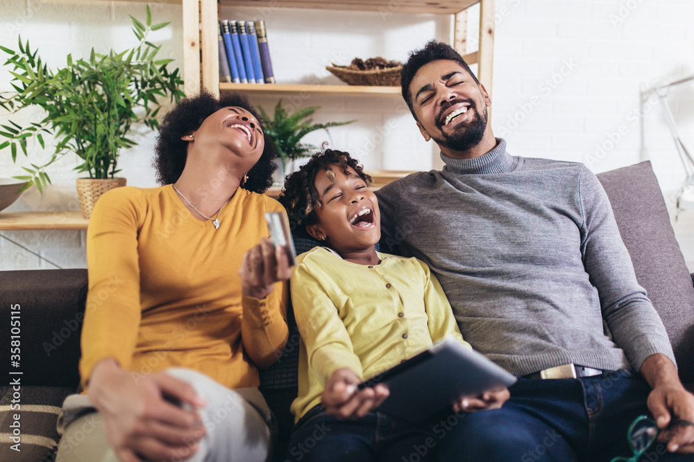 Fototapeta premium Cute little Afro-American girl and her beautiful young parents using digital tablet and doing shopping online while sitting on a sofa at home.