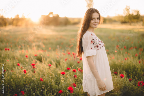 Young happy smile caucasian girl staying at a green red poppy field in the morning. The background of the sun shines. Concept of freedom. Summer. Beautiful young woman walking on field