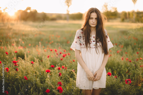 Young happy smile caucasian girl staying at a green red poppy field in the morning. The background of the sun shines. Concept of freedom. Summer. Beautiful young woman walking on field