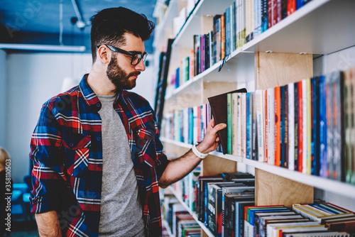 Successful joyful man student holding positive book while standing near shelves in university library