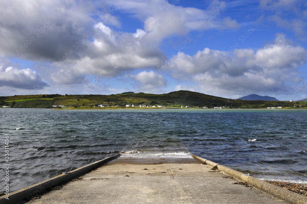 Rampe plongeant dans la mer agitée par le vent sur la côte nord irlandaise, avec des collines et un petit village sur fond de ciel nuageux.