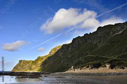 Dunes de sable et herbe verdoyante sur une plage du littoral au nord de l'Irlande. Ciel bleu et nuages blancs contrastant avec les couleurs des falaises.