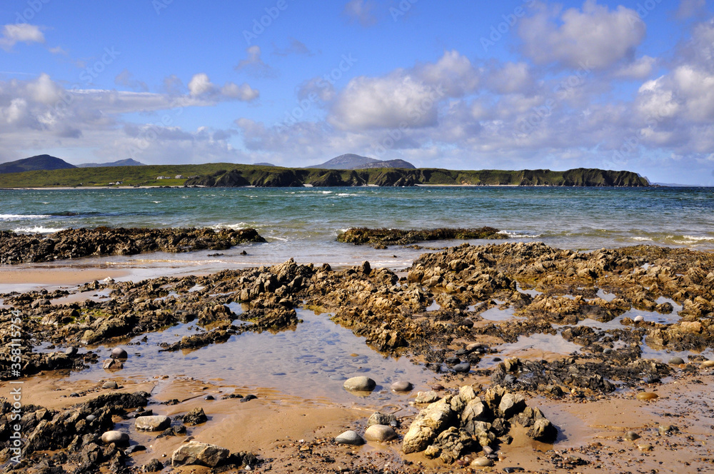 Magnifique plage sauvage donnant sur une mer bleue, verte et turquoise avec rochers et falaises vertes à l'horizon. Ciel bleu et nuages blancs offrant un contraste saisissant.