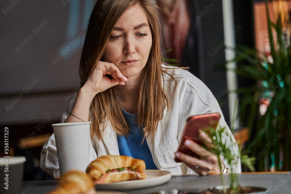Front view of attractive woman wearing glasses using smartphone, drinking coffee in cafe. Female texting and sharing messages on social media, enjoying mobile technology, relaxing in coffee shop.