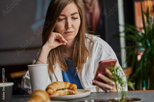 Front view of attractive woman wearing glasses using smartphone  drinking coffee in cafe. Female texting and sharing messages on social media  enjoying mobile technology  relaxing in coffee shop.