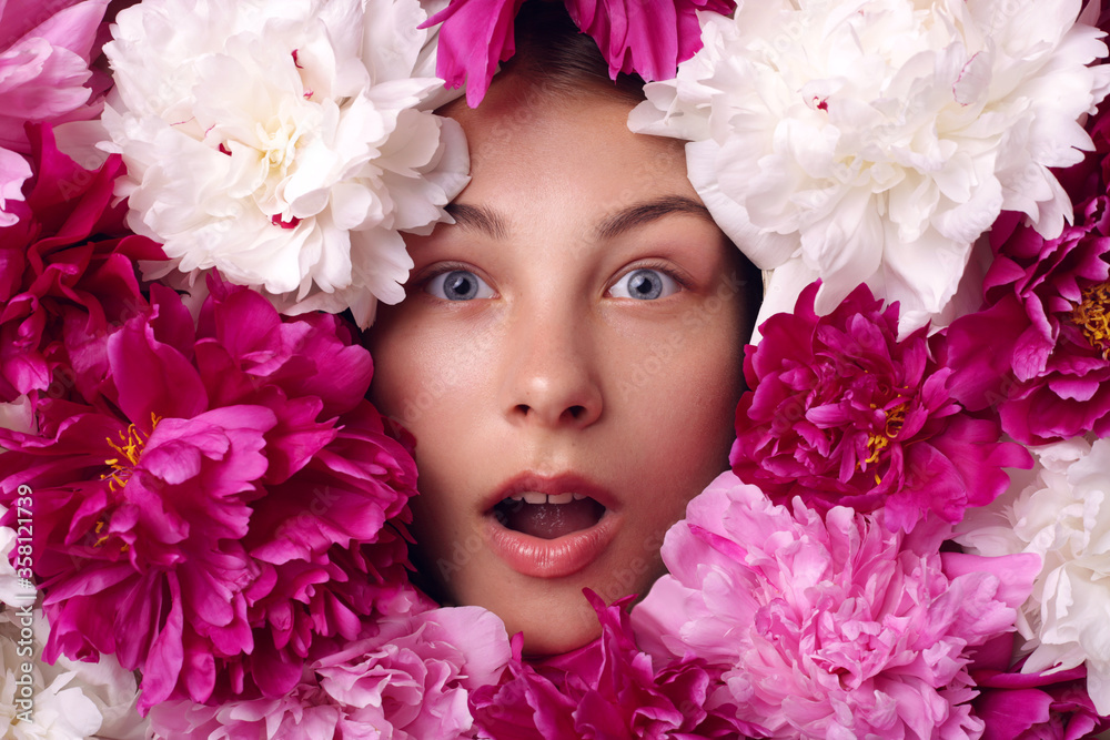 Attractive woman with opened mouth mouth, with astonished facial expression, female with beautiful eyes looks shocked, girl surrounded with white, pink and burgundy peony flowers.