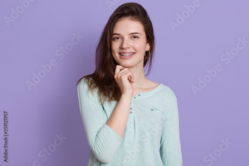 Happy woman with dark hair keeping fist under chin, looks at camera, wears casual attire, poses against lilac background, expressive positive emotions. © sementsova321