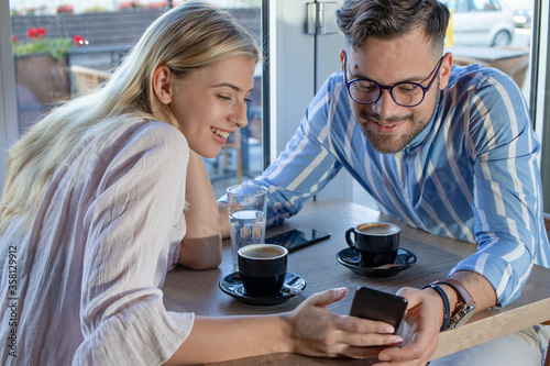 Young romantic couple enjoying a cup of coffee together in a cafe  sitting at the table and talking while scrolling through social media on the smartphone.