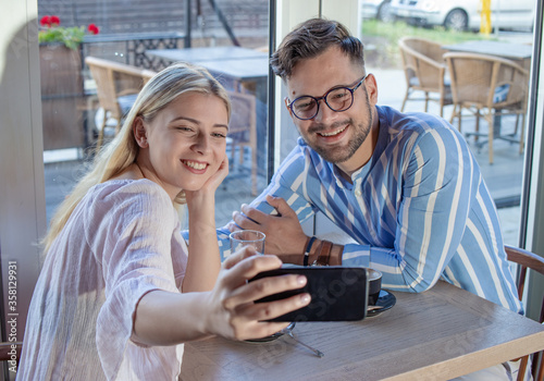 Young romantic couple enjoying a cup of coffee together in a cafe, sitting at the table and taking a selfie with a smartphone.