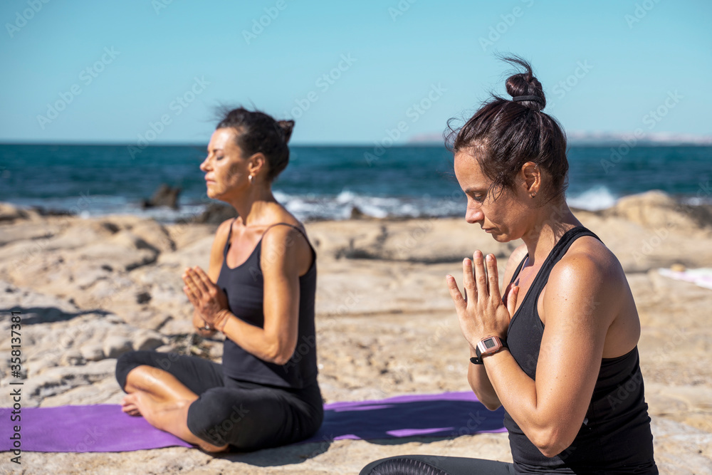 Fototapeta premium Two woman meditating on a rocky beach in sunny day in lotus pose