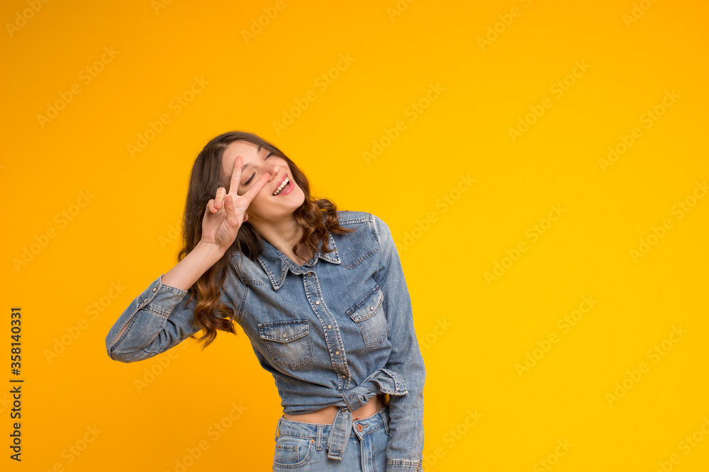 Happy caucasian brunette young woman in denim clothes closed eyes in pleasure,toothy smiling having fun showing peace victory gesture hold disco sign eye tilting head, isolated on yellow background Happy caucasian brunette young woman in denim clothes closed eyes in pleasure,toothy smiling having fun showing peace victory gesture hold disco sign eye tilting head, isolated on yellow background