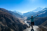 A woman wearing a beanie and blue jumper, spreads her arms wide, breathing deeply the fresh mountain air. Freedom and happiness. Below Manang valley stretches in Himalayas, along Annapurna Circuit.