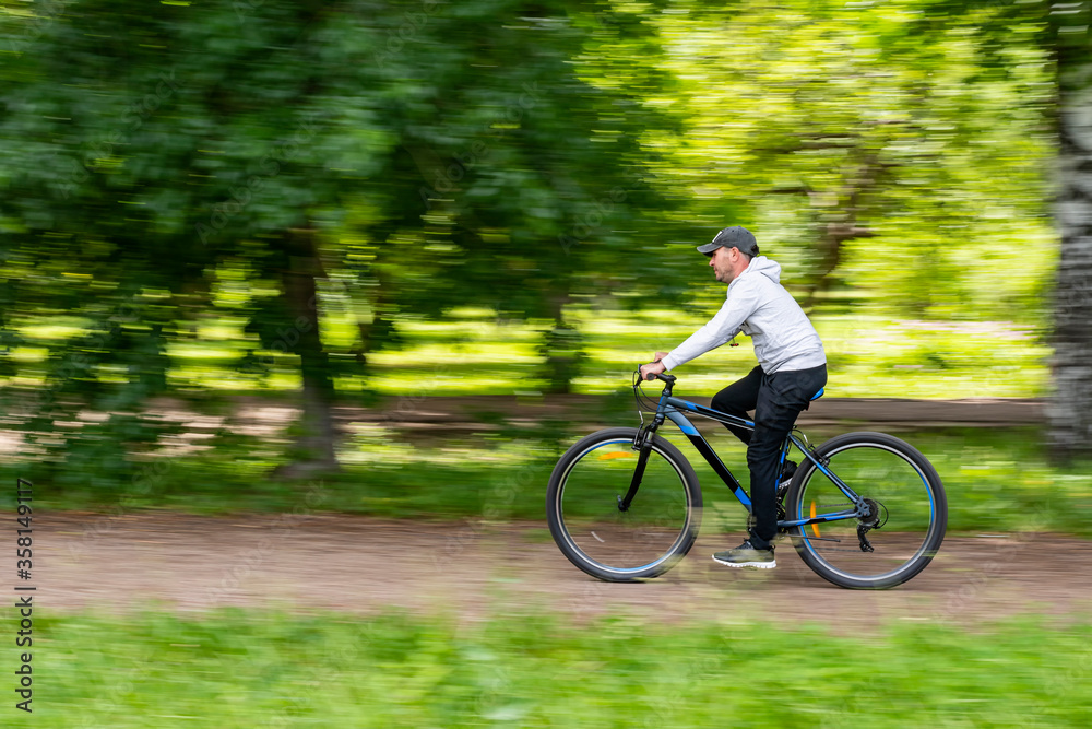 A young guy rides a bicycle at high speed on a treadmill of a school stadium against the background of an apple orchard. Blurred abstract green background.