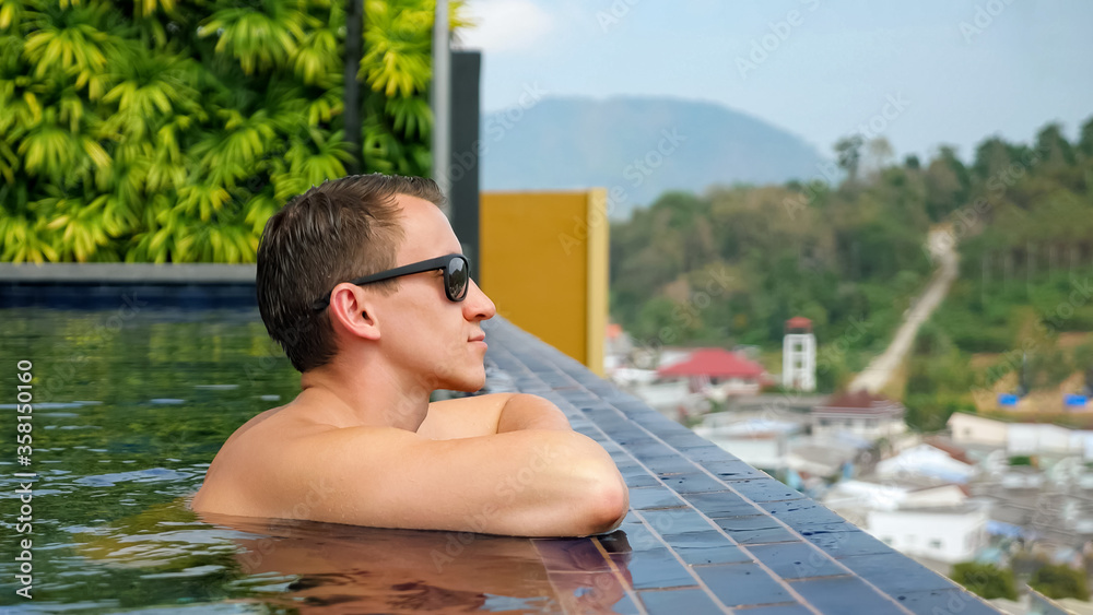 handsome man in sunglasses leans on hotel swimming pool edge and looks at tropical landscape downwards close view
