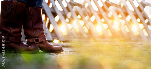 The girl stands on the ranch in cowboy boots and catches the rays of the setting sun.