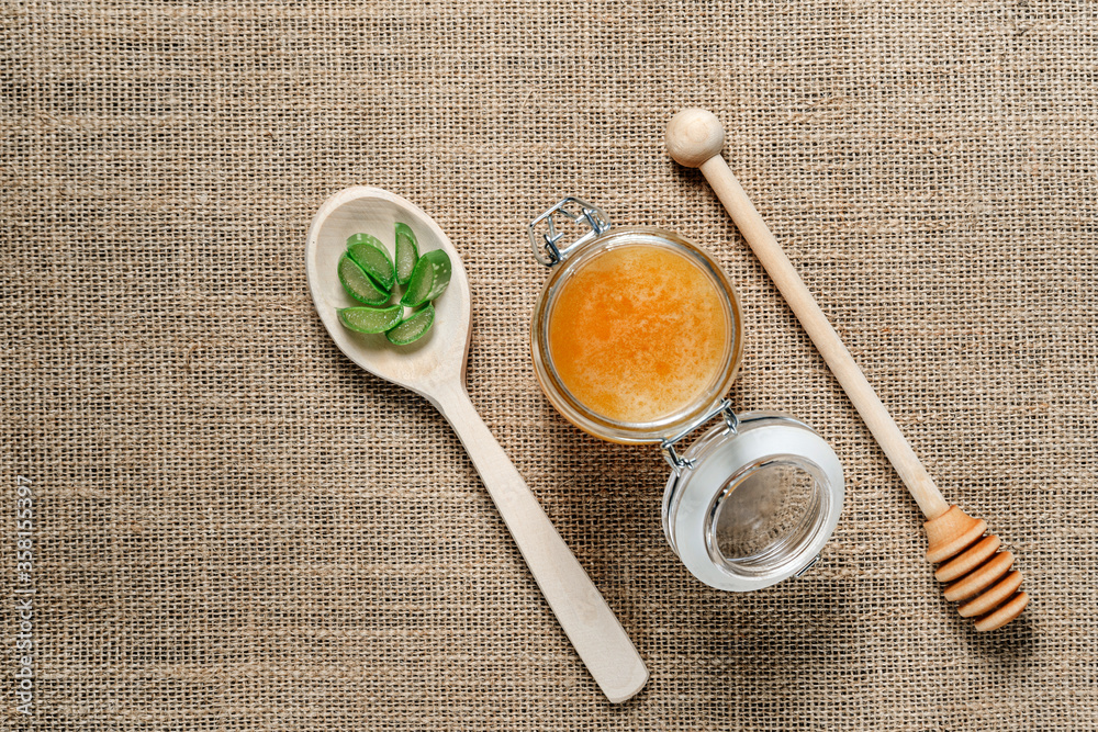 Fototapeta premium Jar of honey, a wooden spoon for honey and a spoon with chopped aloe vera leaves on burlap.