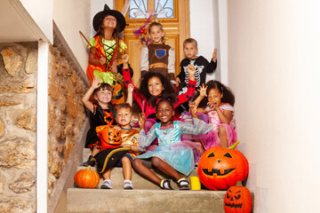 Halloween group kids with spooky gestures sit together boys, girls on the stairs near house door in costumes holding pumpkin, buckets scream and smile