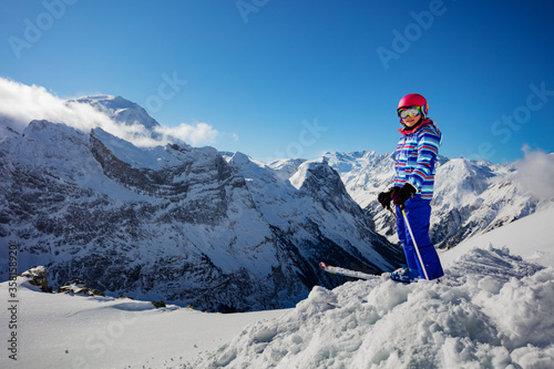 Portrait of a ski girl stand on snow pile in colorful vivid sport clothes, helmet and glasses over Alps mountain tops view