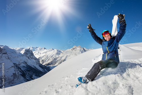 Portrait in the mountains of a girl in blue helmet and ski outfit over the sky sit in snow lifting hands on alpine resort
