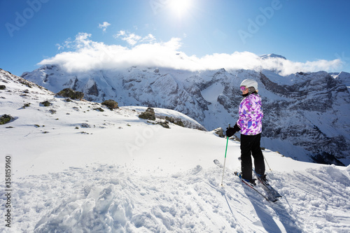 Cute happy teenage girl stand on top of high mountain on ski with winter sport outfit view from behind