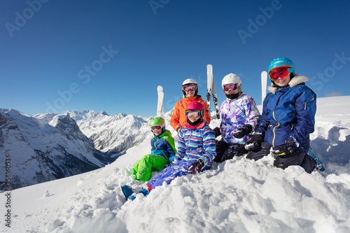 Large group of ski kids sit on the pile of snow over mountain range peaks in colorful sport outfit