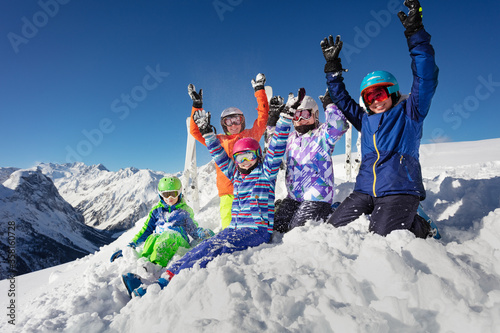 Large group of ski kids sit on the pile of snow over mountain range peaks in colorful sport outfit and rise hands