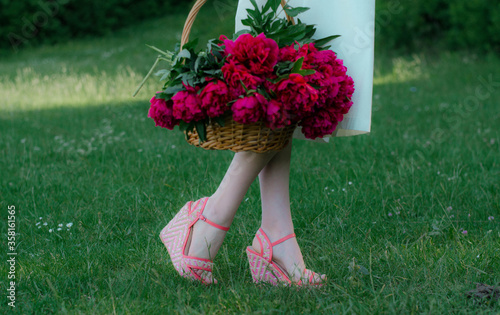 A girl in a white dress stands in green grass in sandals and holds a basket with red peonies